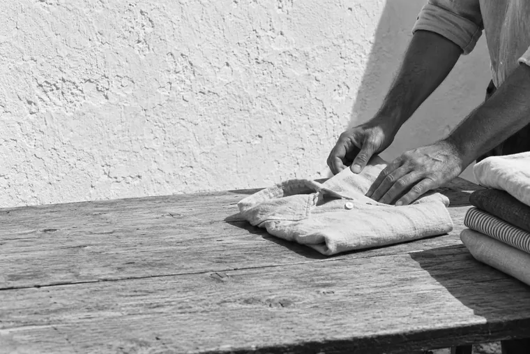 Two hands folding a linen shirt on a bare wooden table, a stack of pressed clothes at the edge of the frame, a limewashed wall in full sun