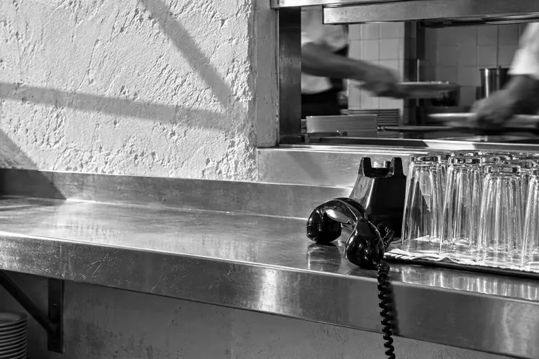 Le passe d’un restaurant en plein service : des verres alignés au premier plan, un combiné téléphonique décroché posé sur l’inox